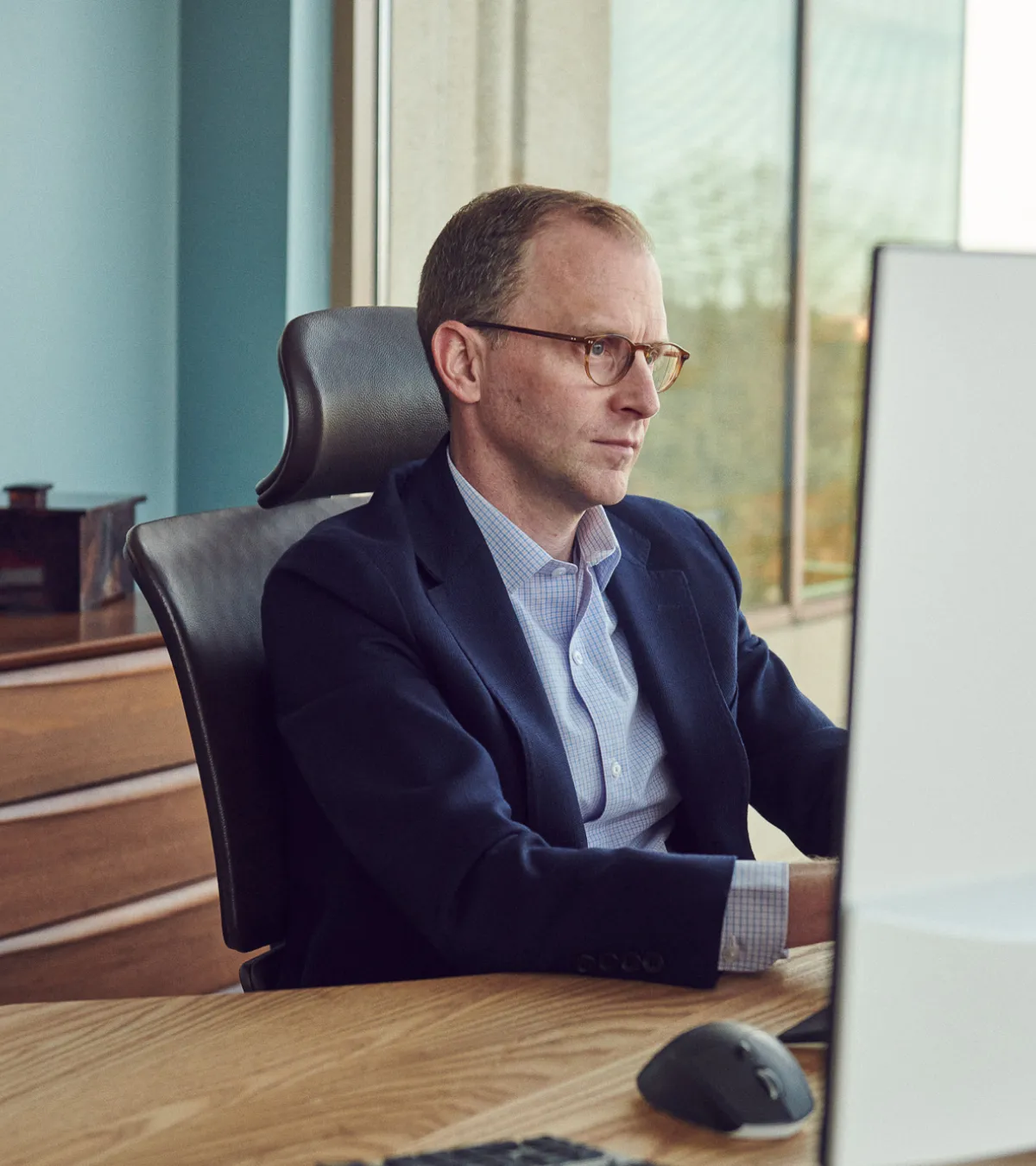 Man in glasses and blazer focused on working at a computer in a modern office.