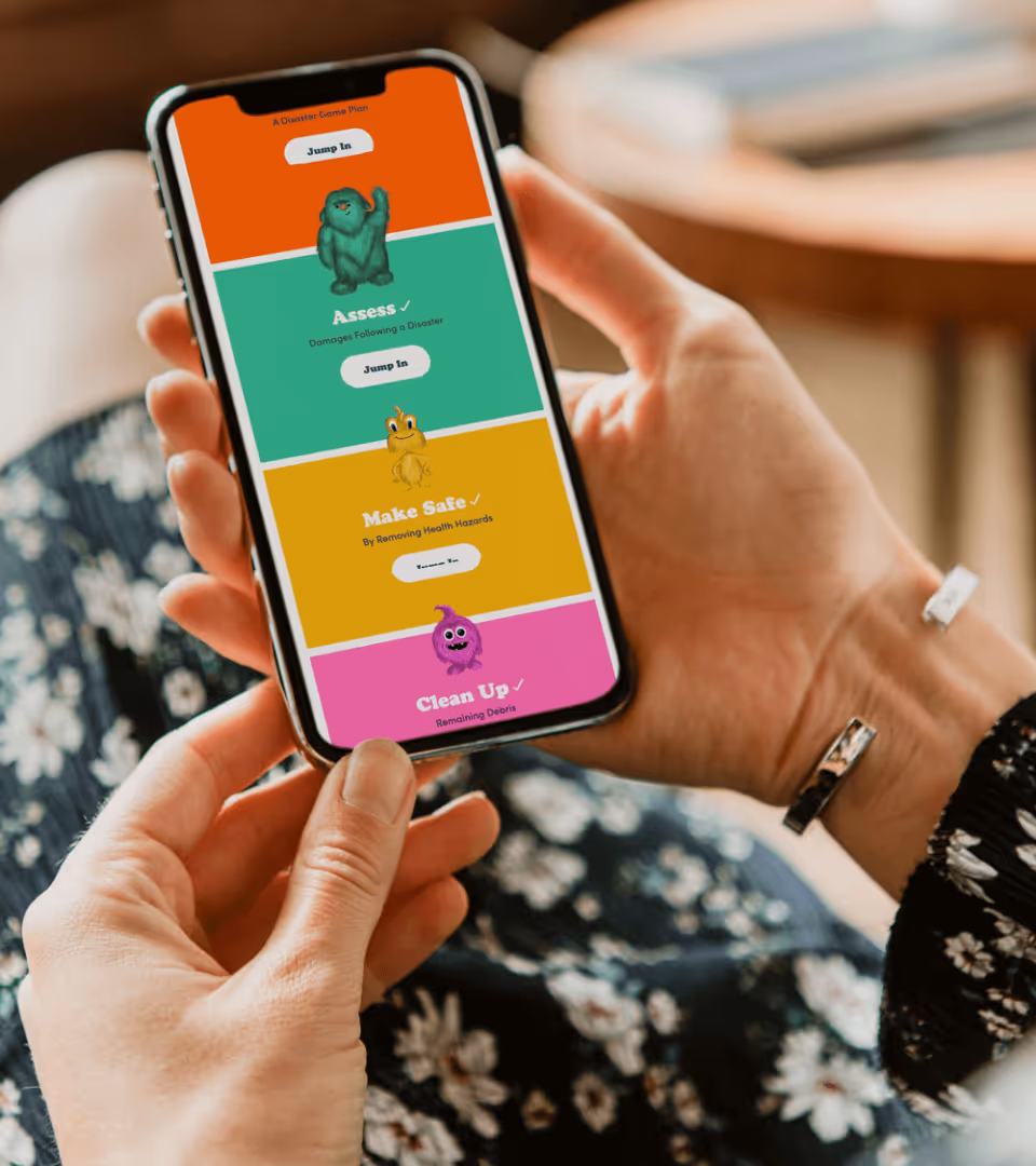 Person holding a smartphone displaying a colorful disaster preparedness app with options: Assess, Make Safe, and Clean Up.