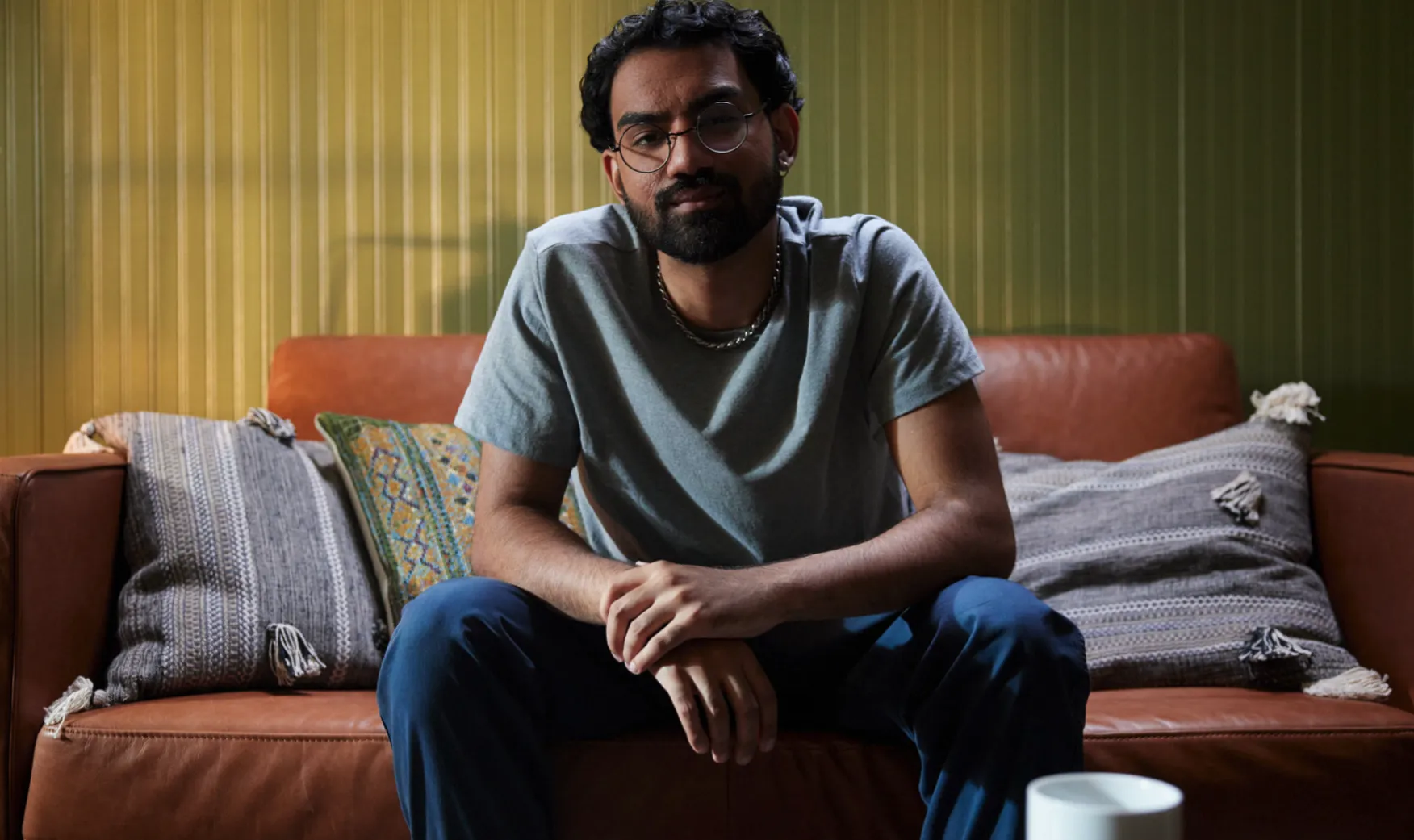 Man with glasses, a beard, and a gray shirt sitting on a brown leather couch with patterned pillows against a green paneled wall.