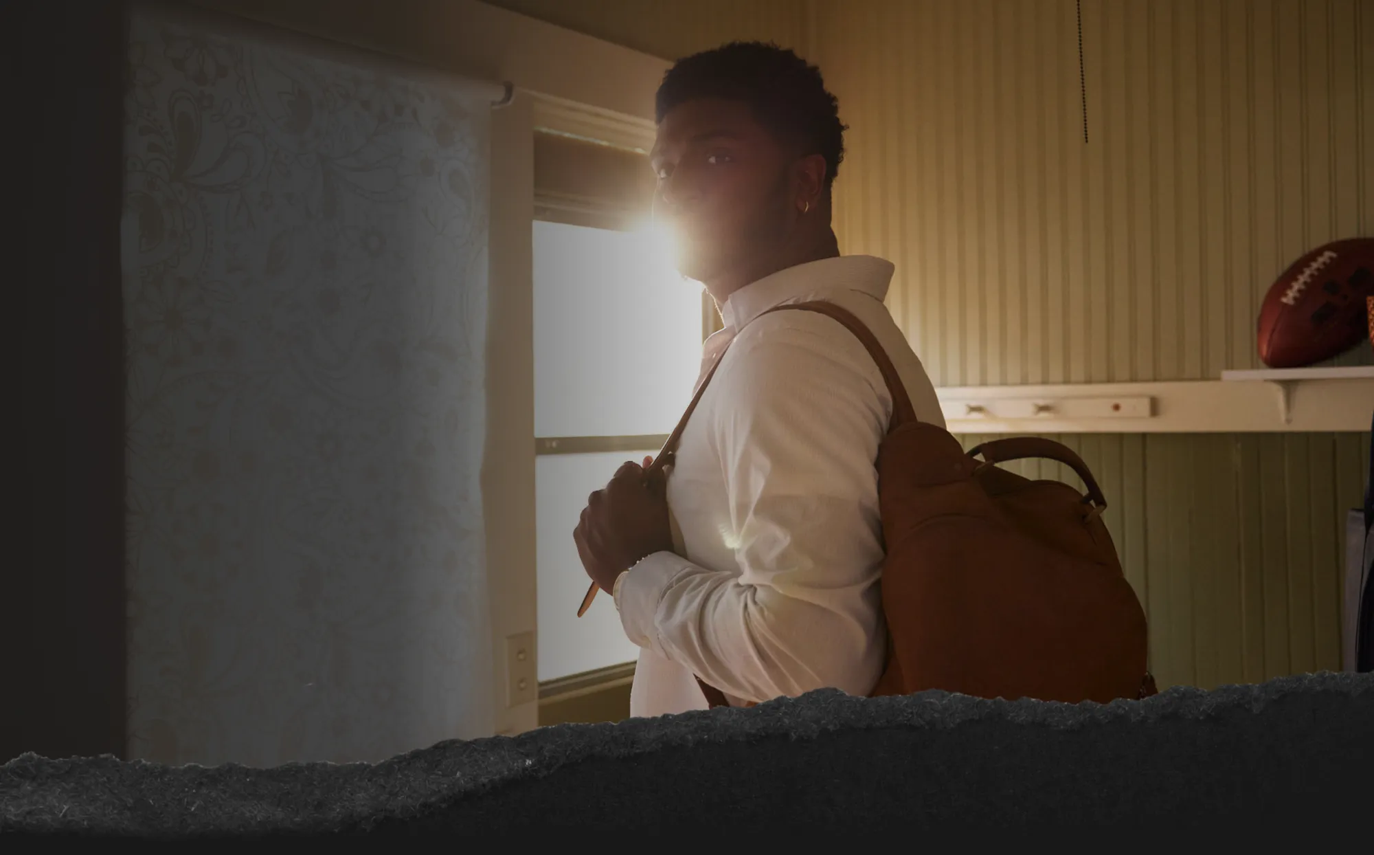 Man wearing a white shirt and carrying a brown backpack, standing indoors with sunlight shining through a window behind him.