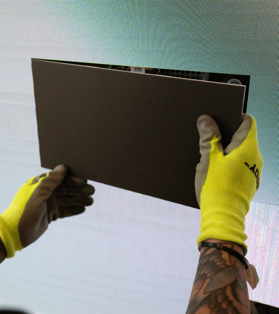 Person wearing yellow gloves and a tattooed arm installing or holding a dark rectangular panel against an LED screen.