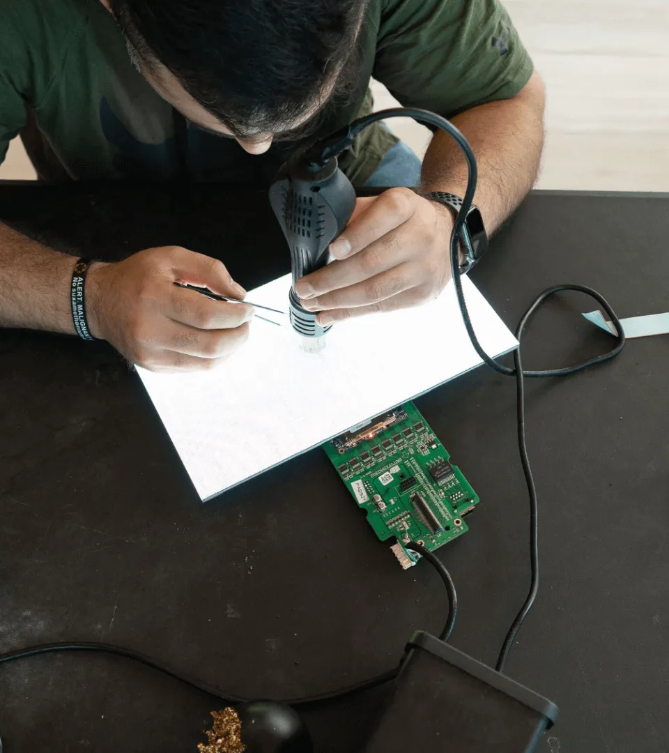 Person inspecting a green circuit board using a magnifying glass with a light source on a white illuminated surface.