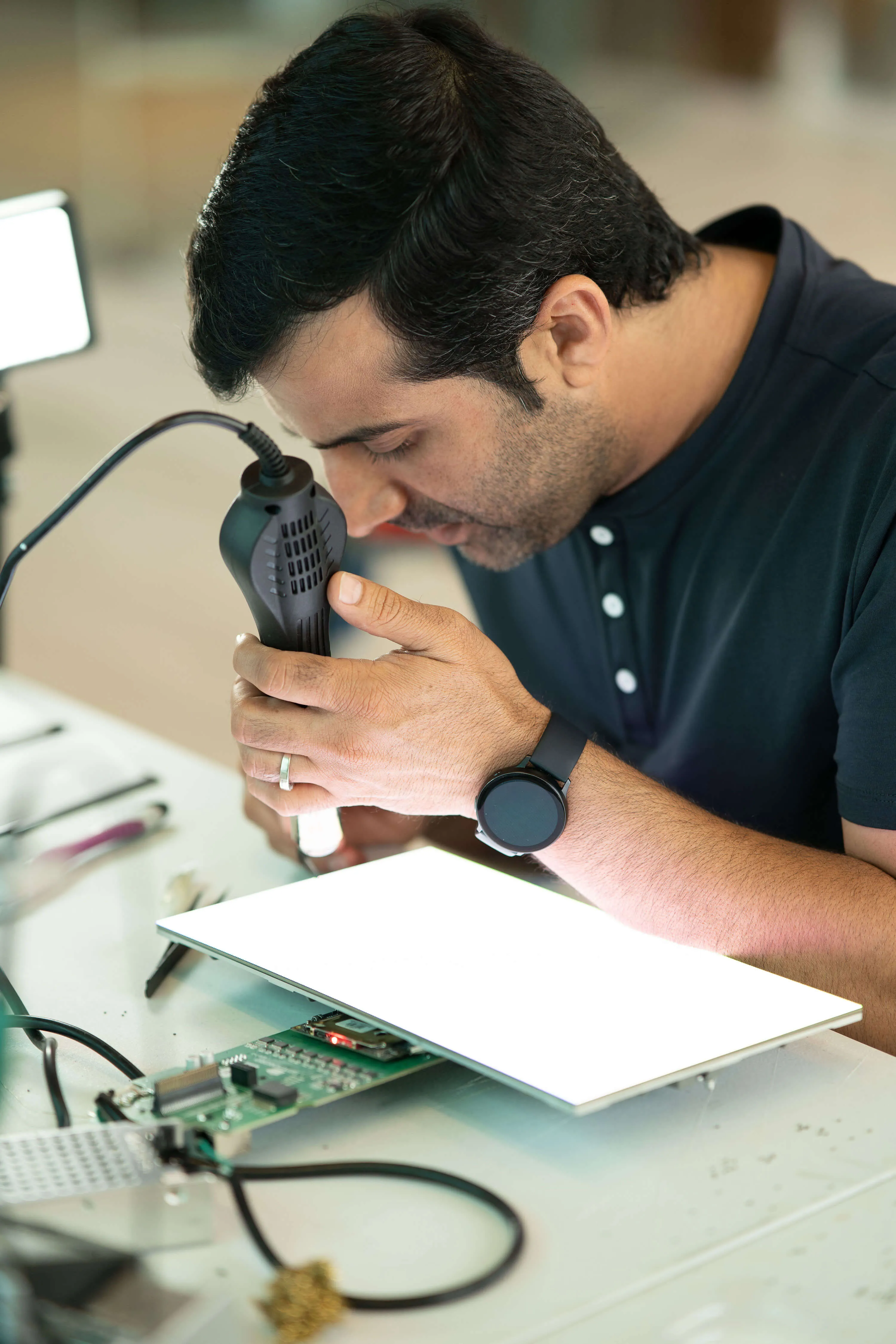 Man inspecting an electronic circuit board using a handheld magnifying tool with an illuminated light panel.