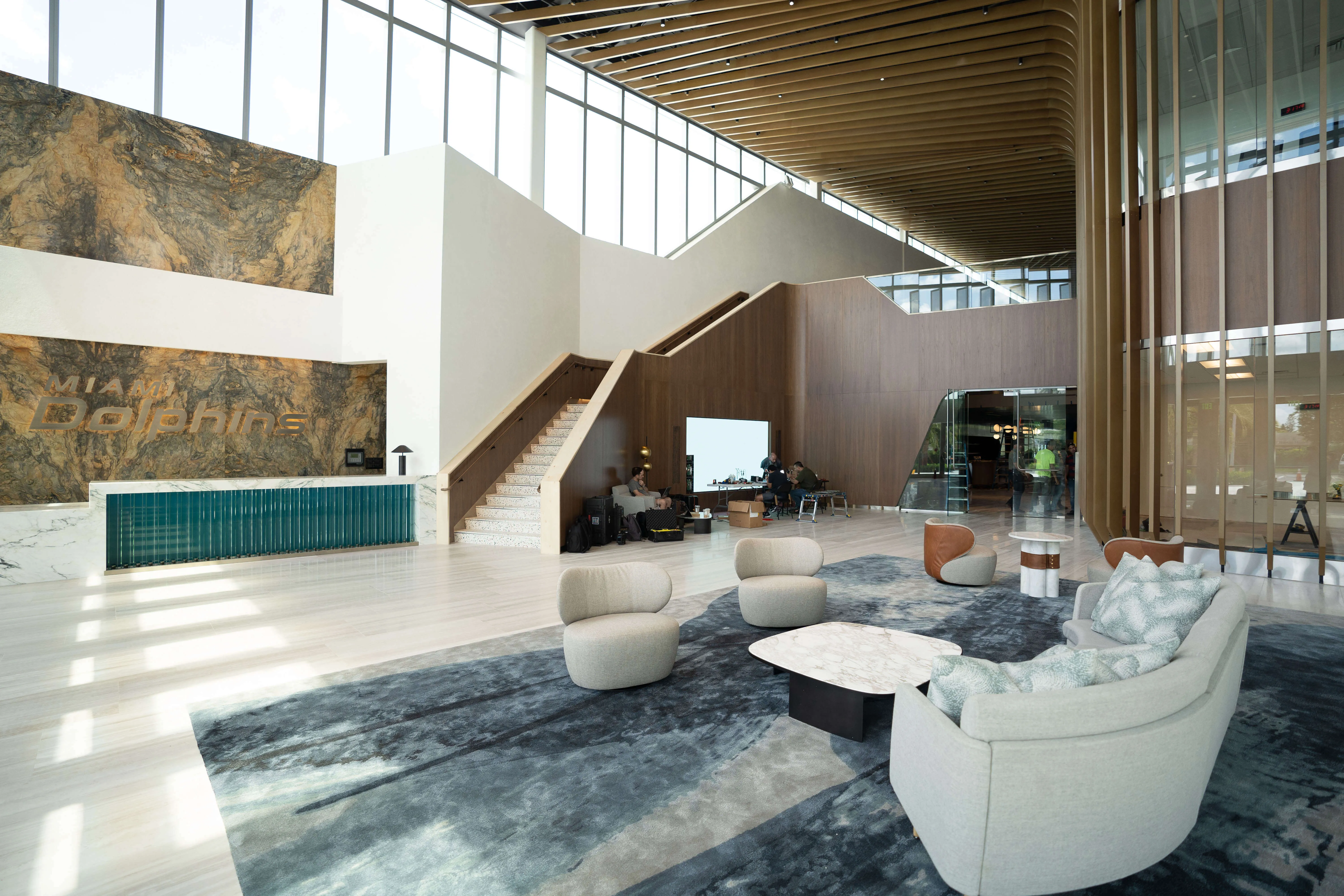 Spacious modern lobby with large windows, light wood flooring, a blue-gray rug, seating area with gray chairs and couch, and a reception desk with Miami Dolphins logo on stone wall.