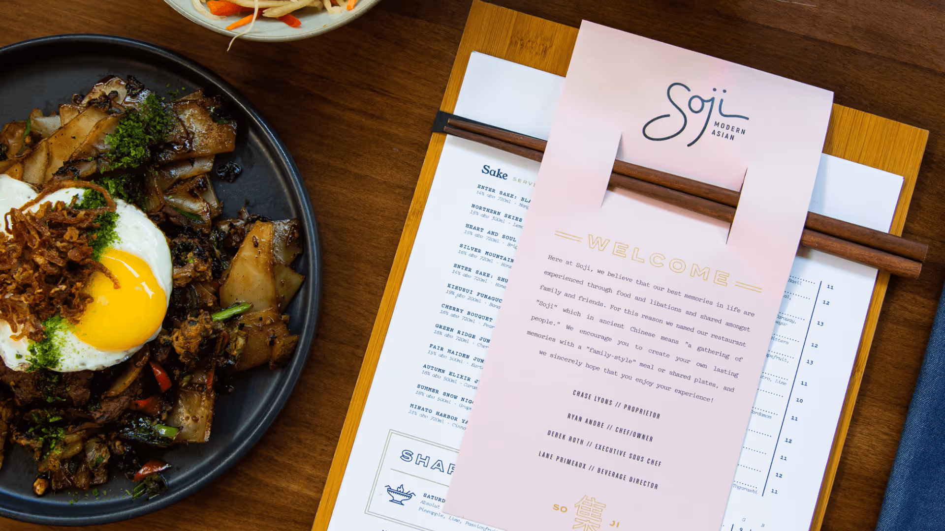 Plate of Asian stir-fried noodles topped with a fried egg and green herbs beside a restaurant menu with chopsticks.
