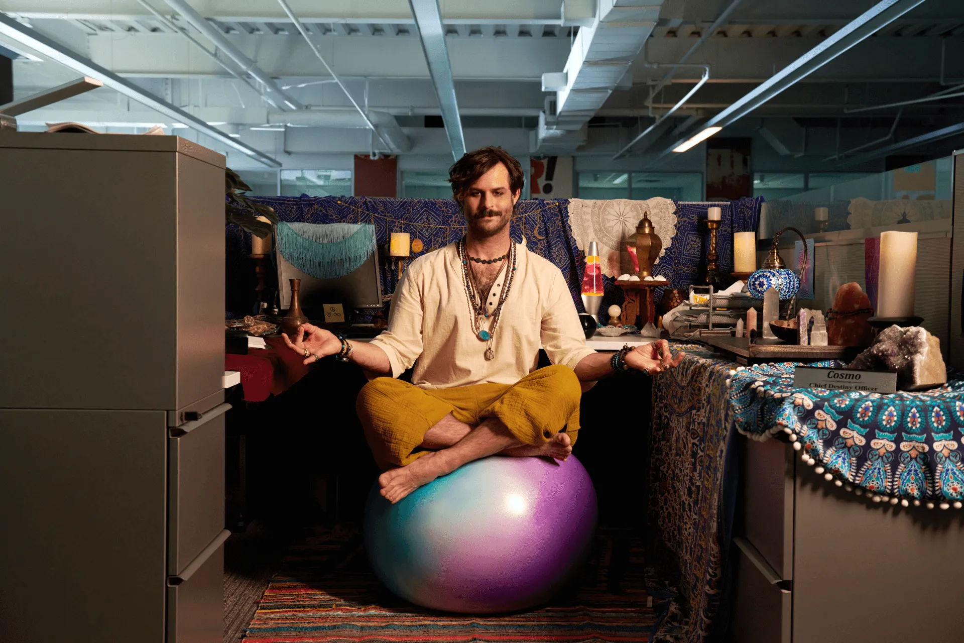 Man in mustard pants and cream shirt sitting cross-legged on a colorful exercise ball meditating in an office cubicle decorated with crystals, candles, and patterned fabrics.