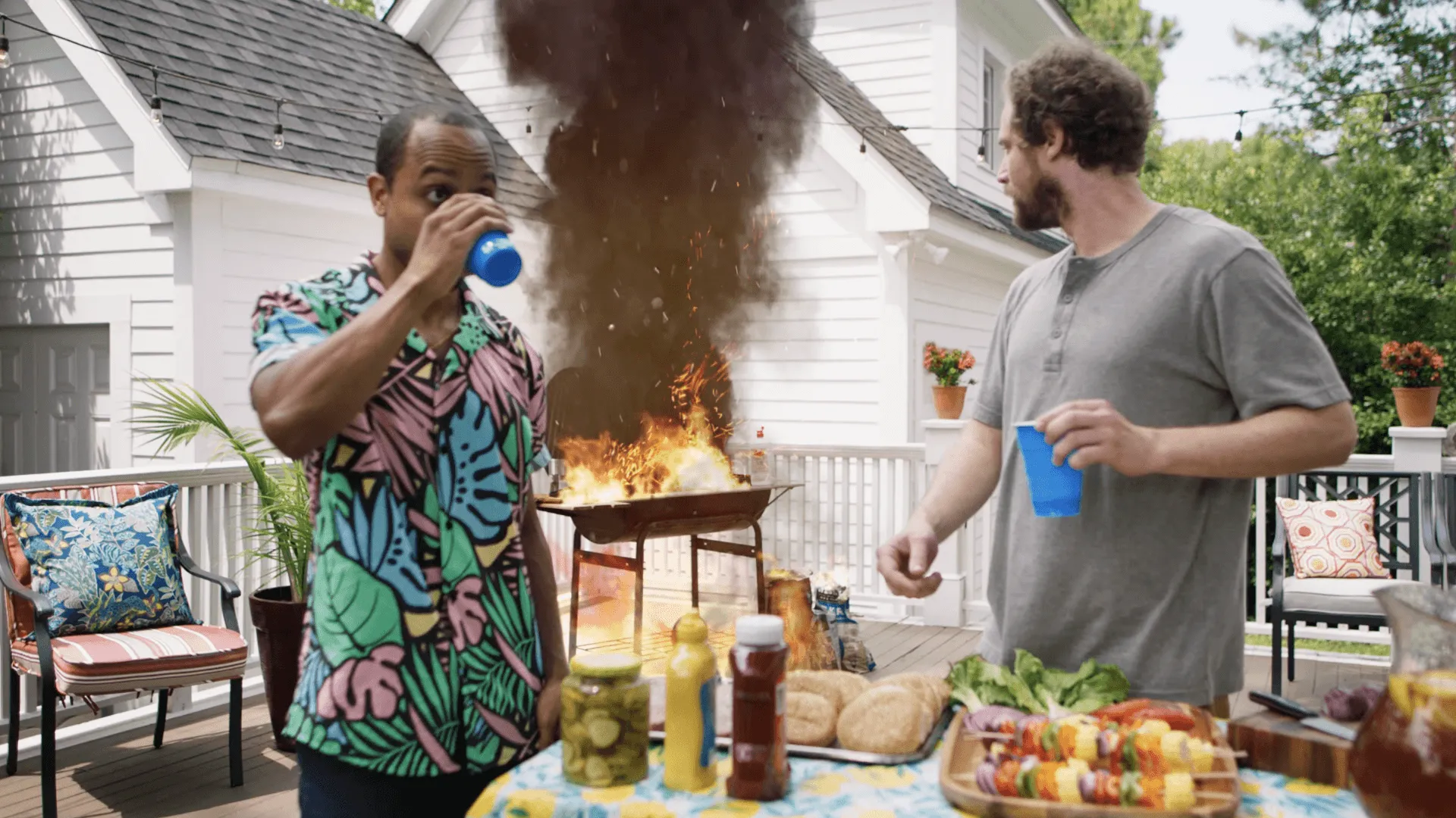 Two men stand on a deck near a barbecue grill that is on fire with thick black smoke, one drinking from a blue cup and the other holding a blue cup looking at the fire.