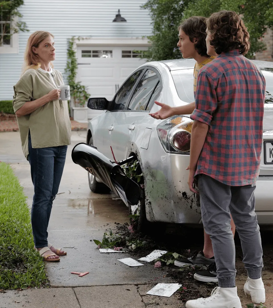 Two teenage boys explaining to a woman holding a mug beside a damaged silver car with a broken mailbox and scattered debris.