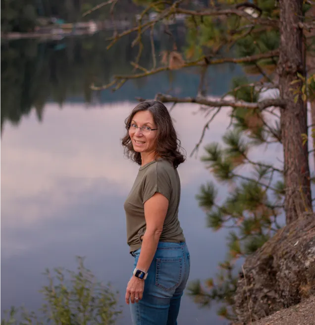 Women Roam founder, Patti Hinman, by a lakeside with trees, smiling and looking back over her shoulder.