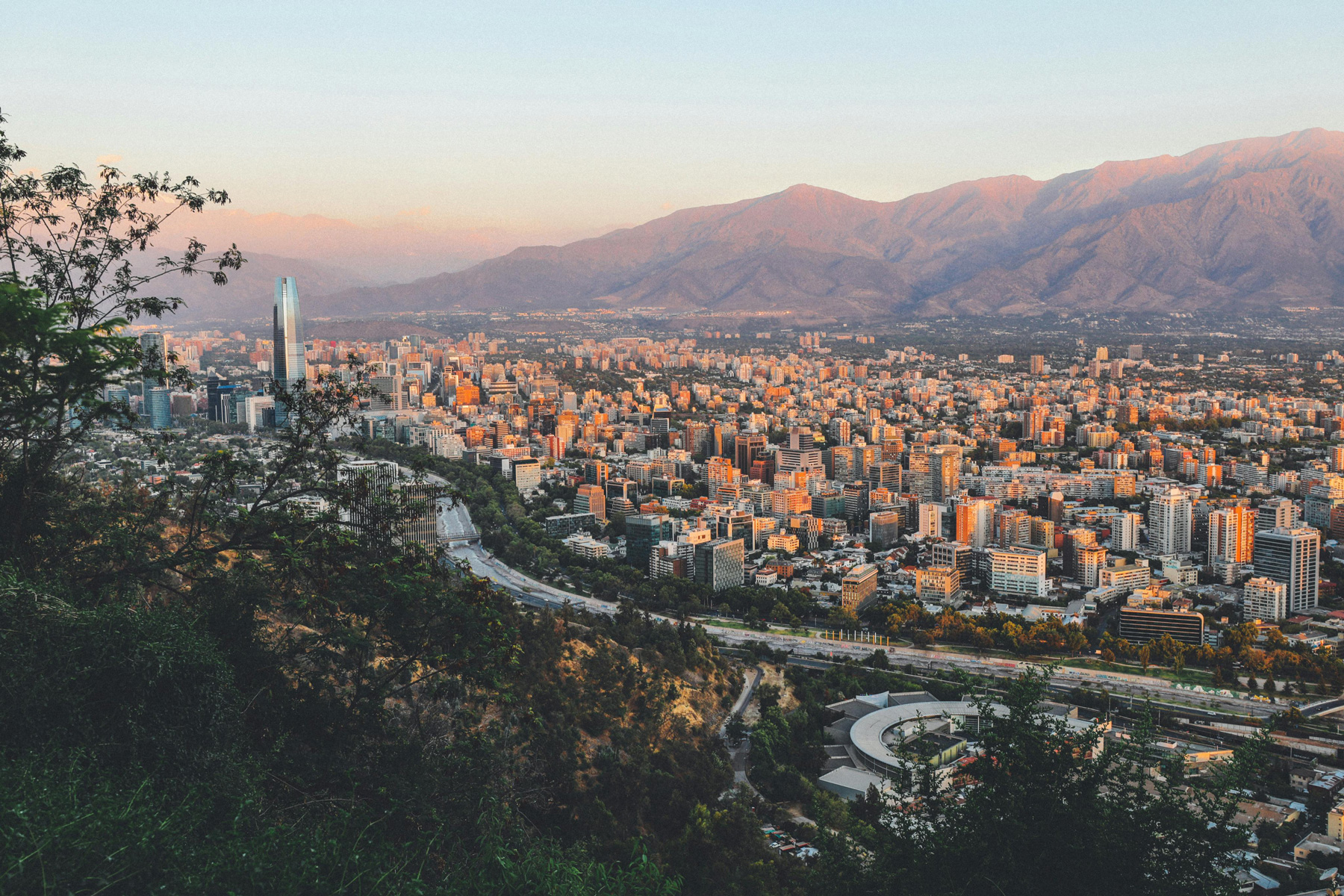 Panoramic view of a cityscape with tall buildings, mountains in the background, and foreground trees during sunset.