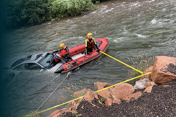 Swiftwater rescue team securing a red inflatable boat beside a partially submerged car.