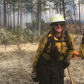 Smiling firefighter in yellow protective gear standing in a forest during wildfire operations.