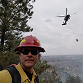 Firefighter wearing a red helmet with a helicopter performing an aerial water drop in the background. 