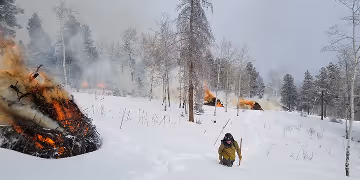 Firefighter monitoring burning brush piles in a snowy forest during controlled burns.