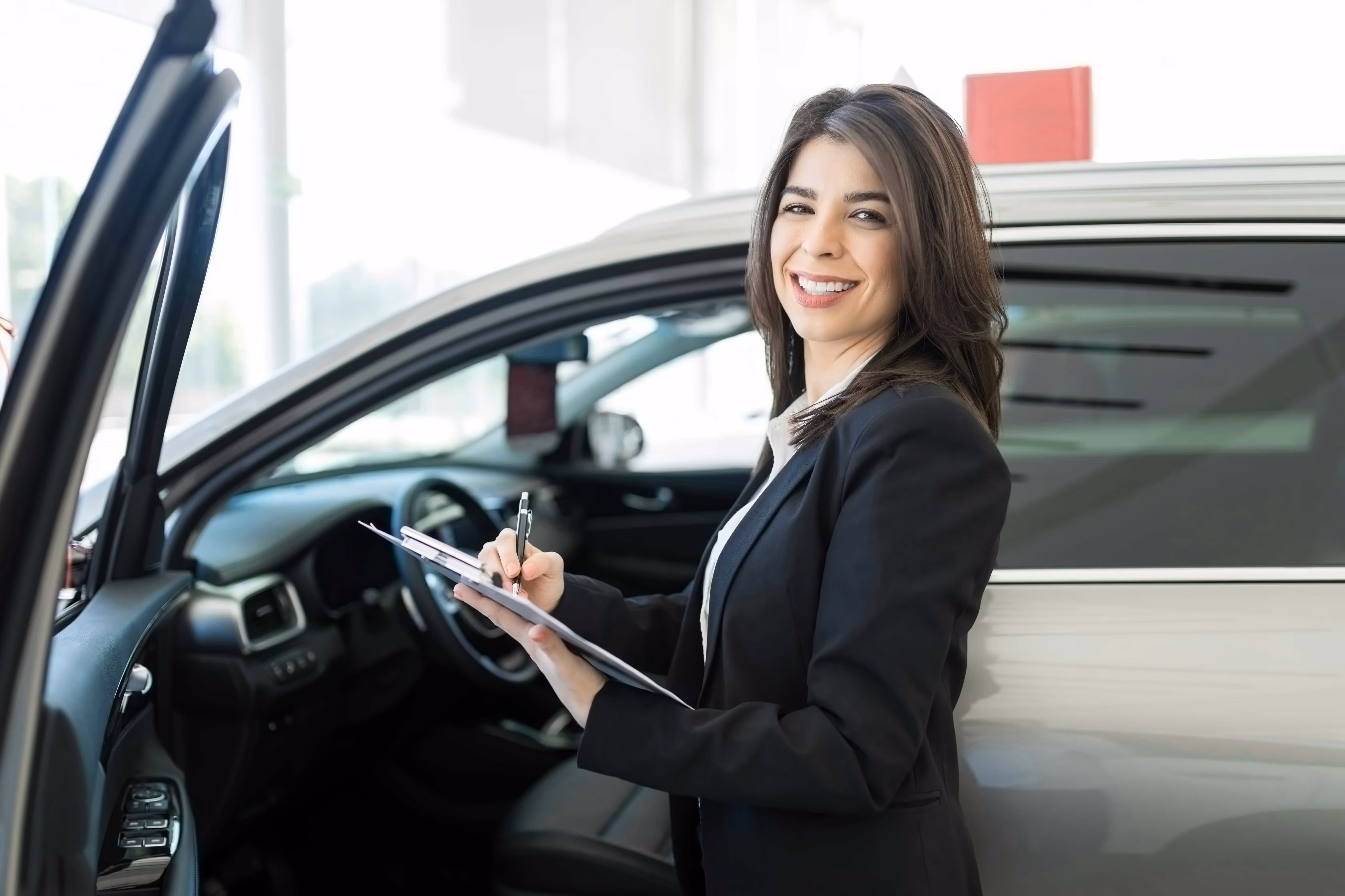 Woman standing beside a car holding a clipboard and smiling at the camera.