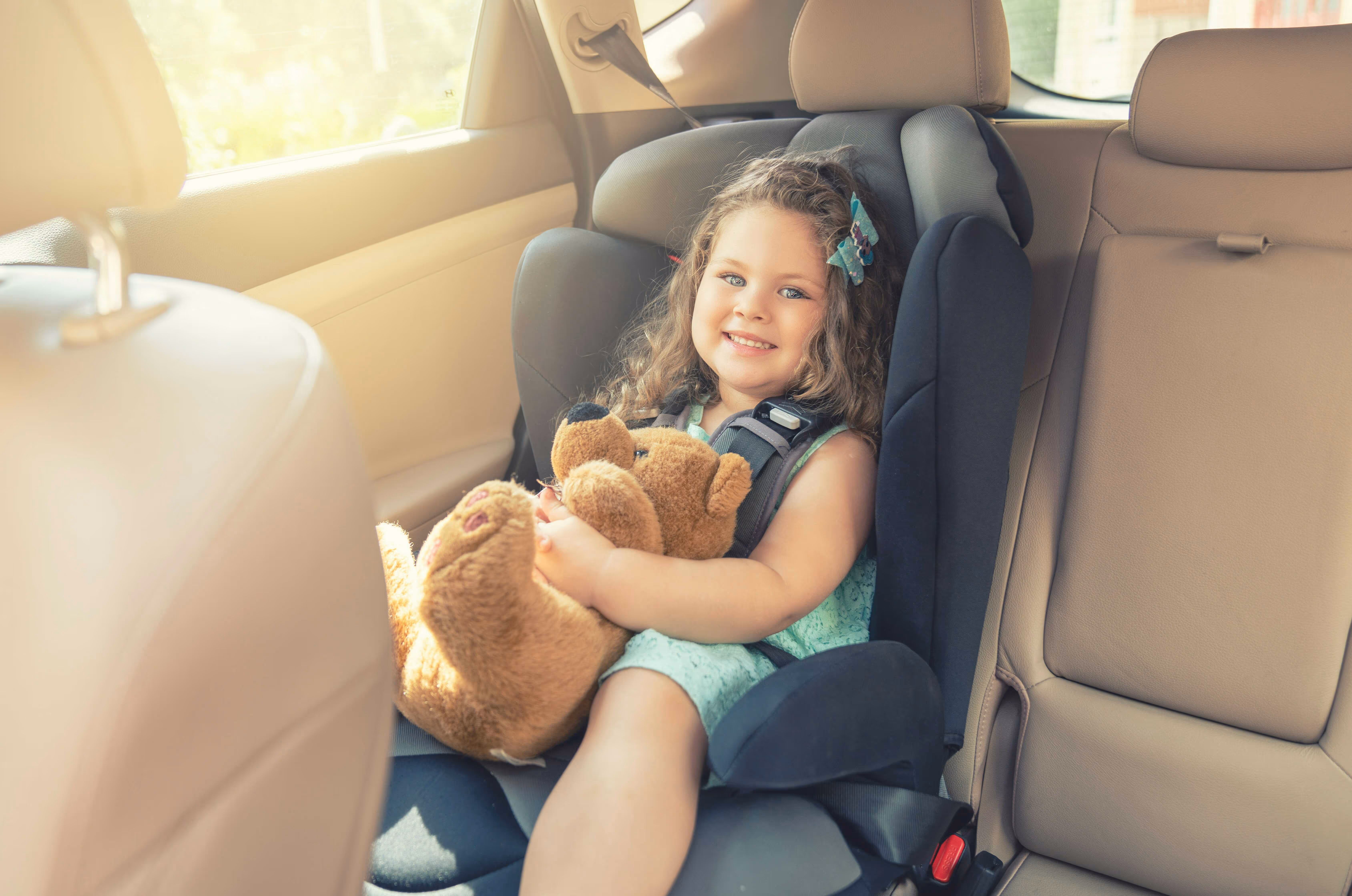 Young girl smiling in a car seat holding a teddy bear inside a vehicle.