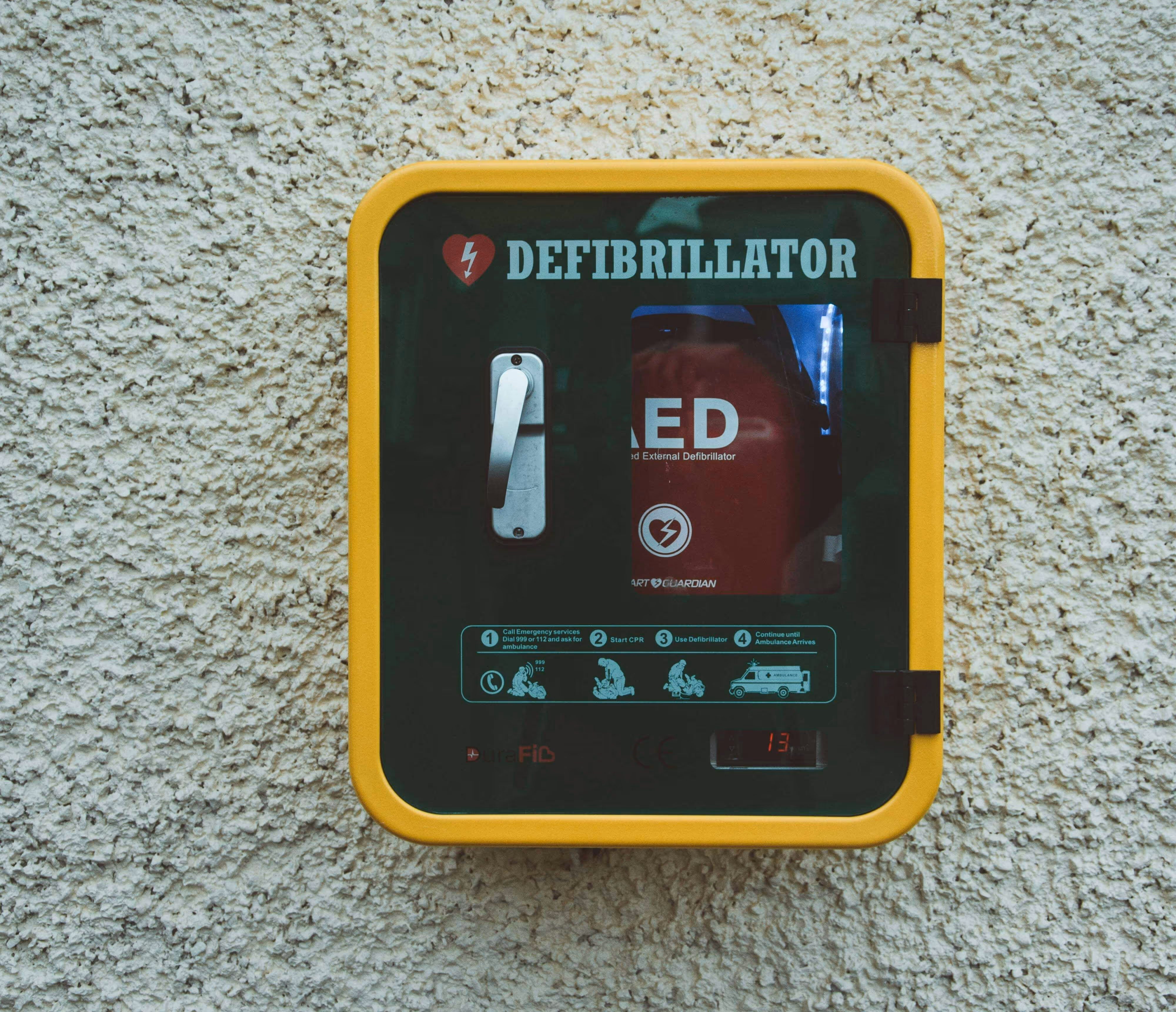 Wall-mounted defibrillator unit in a yellow and green case for emergency use.