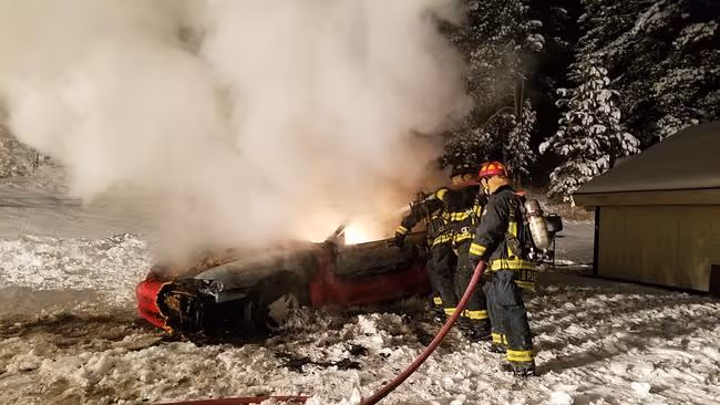 Firefighters spraying water on a burning vehicle surrounded by snow.
