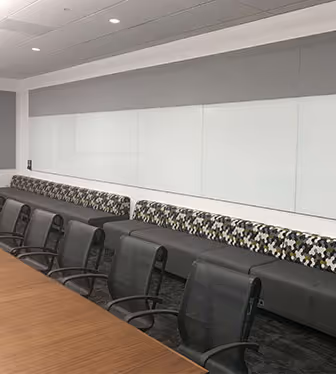 Modern conference room with a long wooden table, mesh office chairs, a cushioned bench with patterned upholstery, and large whiteboards on the wall.