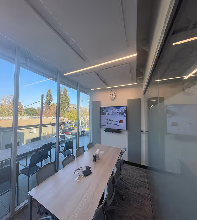 Small modern office meeting room with a wooden table, chairs, wall-mounted TV, clock, and large glass windows overlooking an outdoor area.