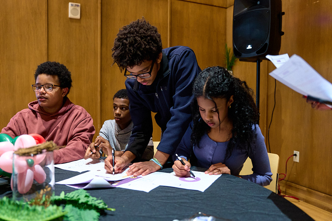 Four young people writing or drawing on papers at a table with a balloon flower decoration in a wood-paneled room.