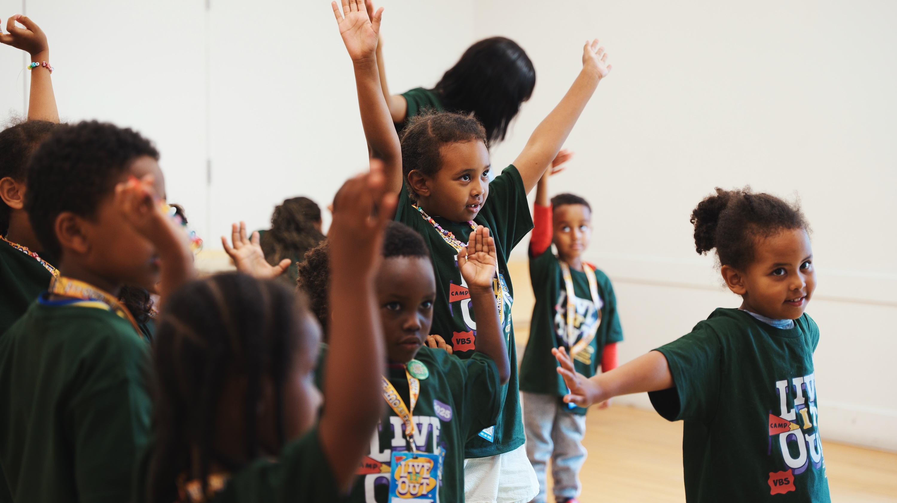 Group of children in green camp shirts raising their hands indoors during an activity.
