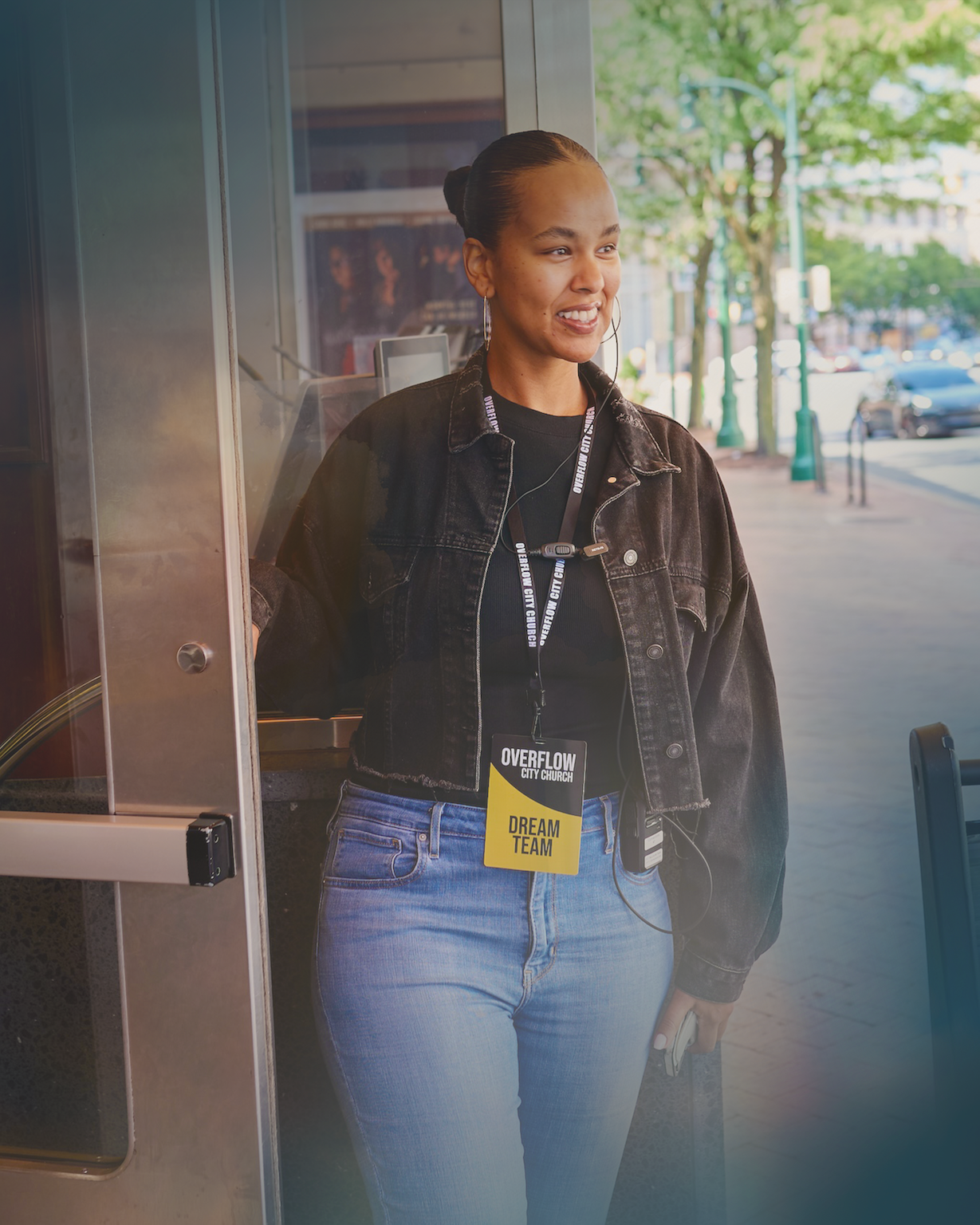 Smiling woman in a black jacket and blue jeans holding a door open, wearing a badge reading 'Overflow City Church Dream Team'.