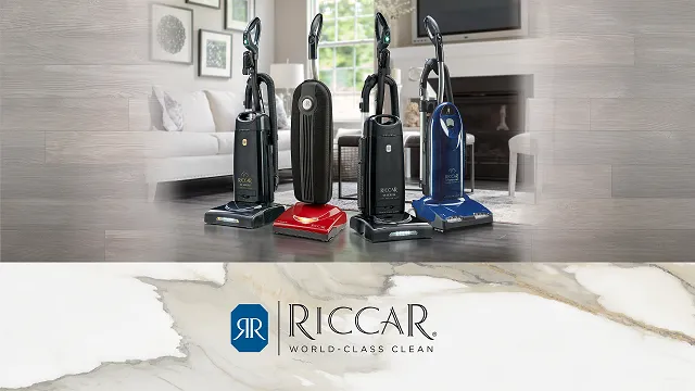 Four upright Riccar vacuum cleaners in different colors displayed in a modern living room, with Riccar logo and slogan 'World-Class Clean' below on a marble background.