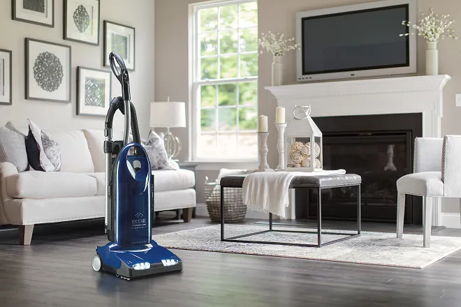 Blue upright vacuum cleaner standing on a wooden floor in a modern living room with white sofas, a coffee table with decor, and a fireplace with a TV above it.
