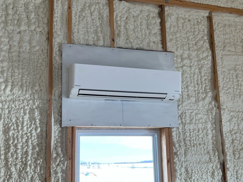 A white air conditioning unit is mounted on a wall with visible wooden studs and foam insulation. 