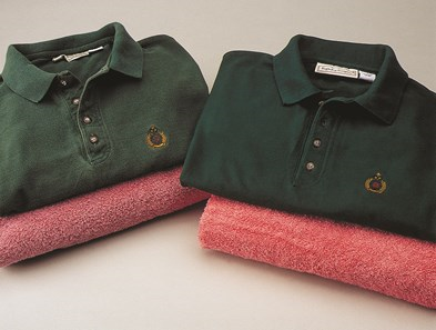 Two green polo shirts folded on top of faded pink towels, showing a before and after contrast in fabric brightness.