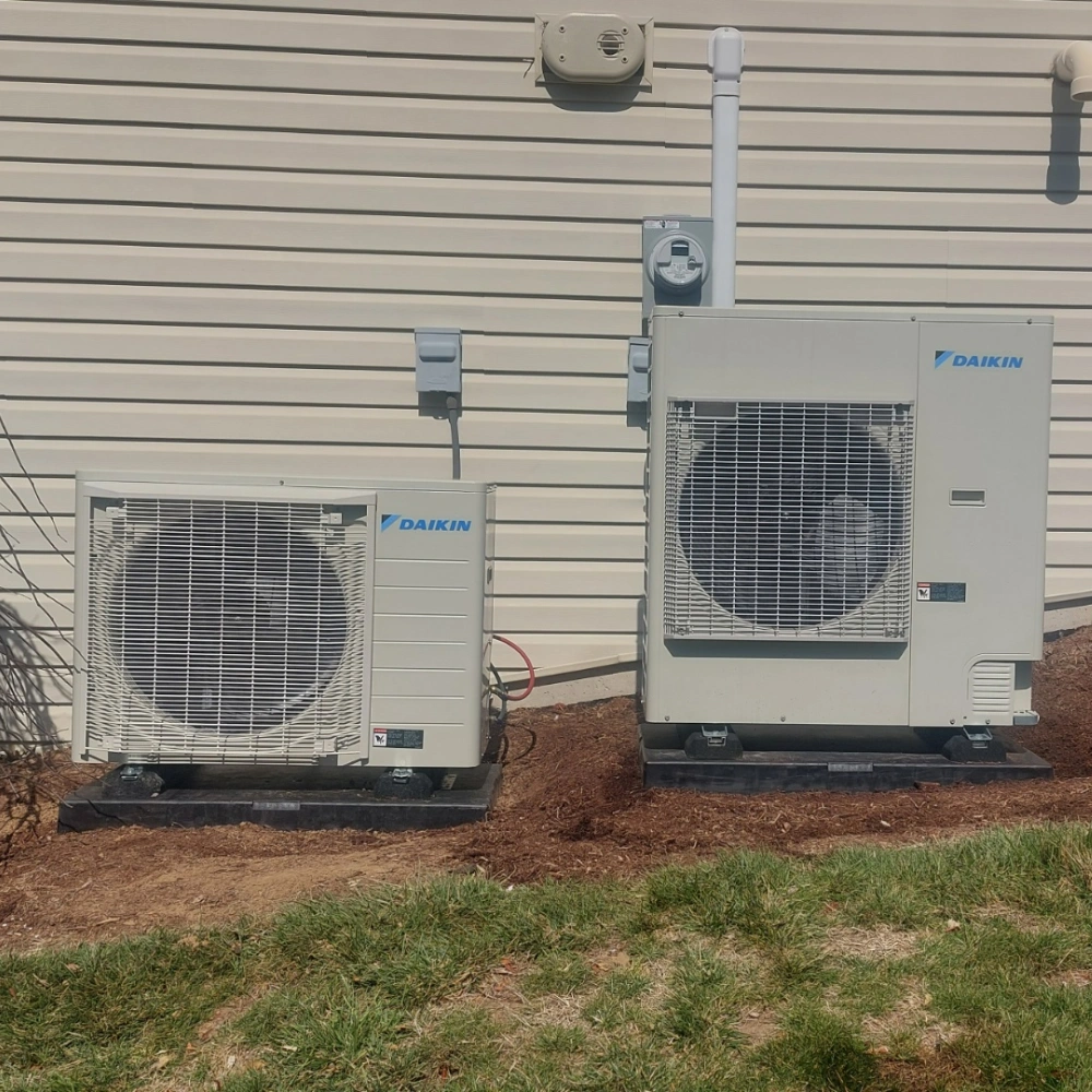 Two outdoor Daikin HVAC units, varying in size, are placed on a concrete area against a beige siding wall. 