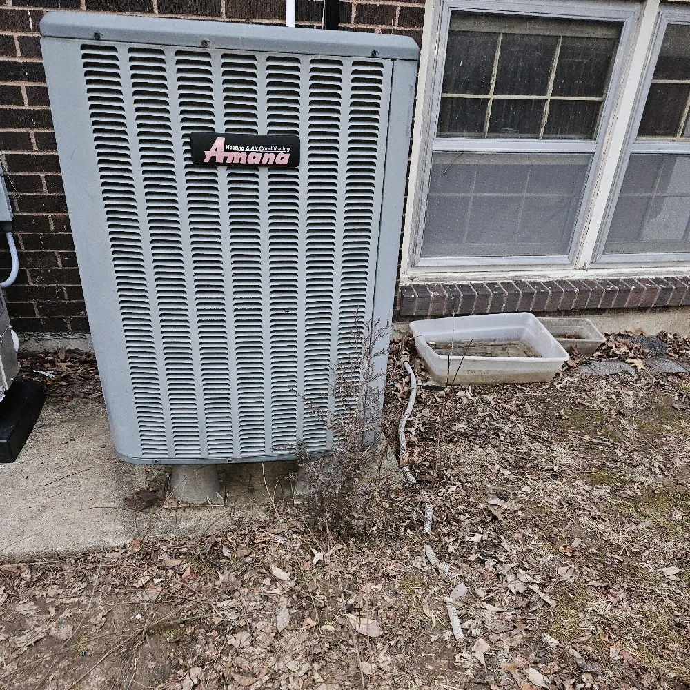 A large gray air conditioning unit labeled "Amana" sits on a concrete pad beside a brick building. 