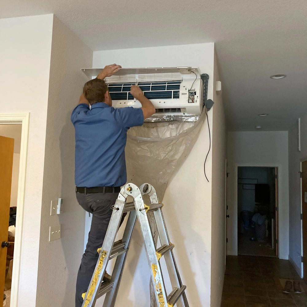 A technician in a blue shirt on a ladder repairs a wall-mounted air conditioner. 