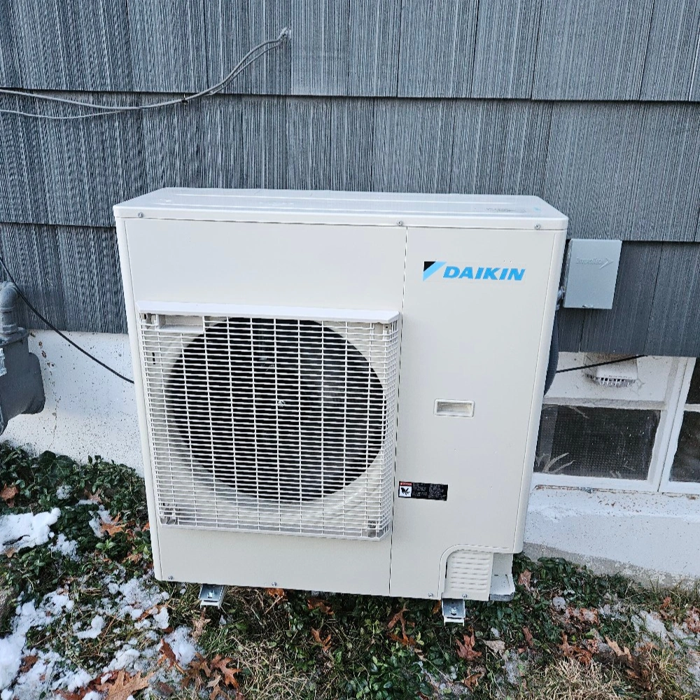 Outdoor Daikin HVAC unit installed beside a gray building with snow patches on the ground. 