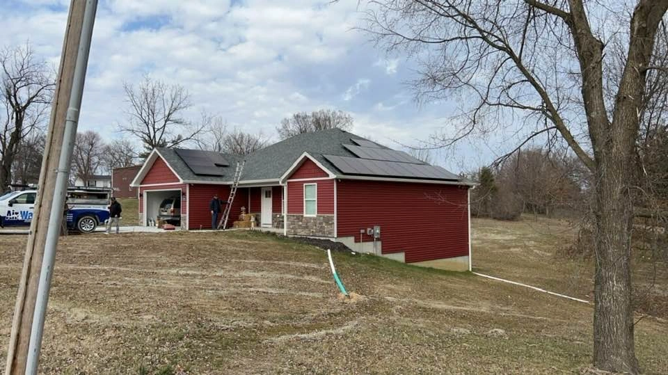 A red house with solar panels on the roof, surrounded by bare trees and a cloudy sky.