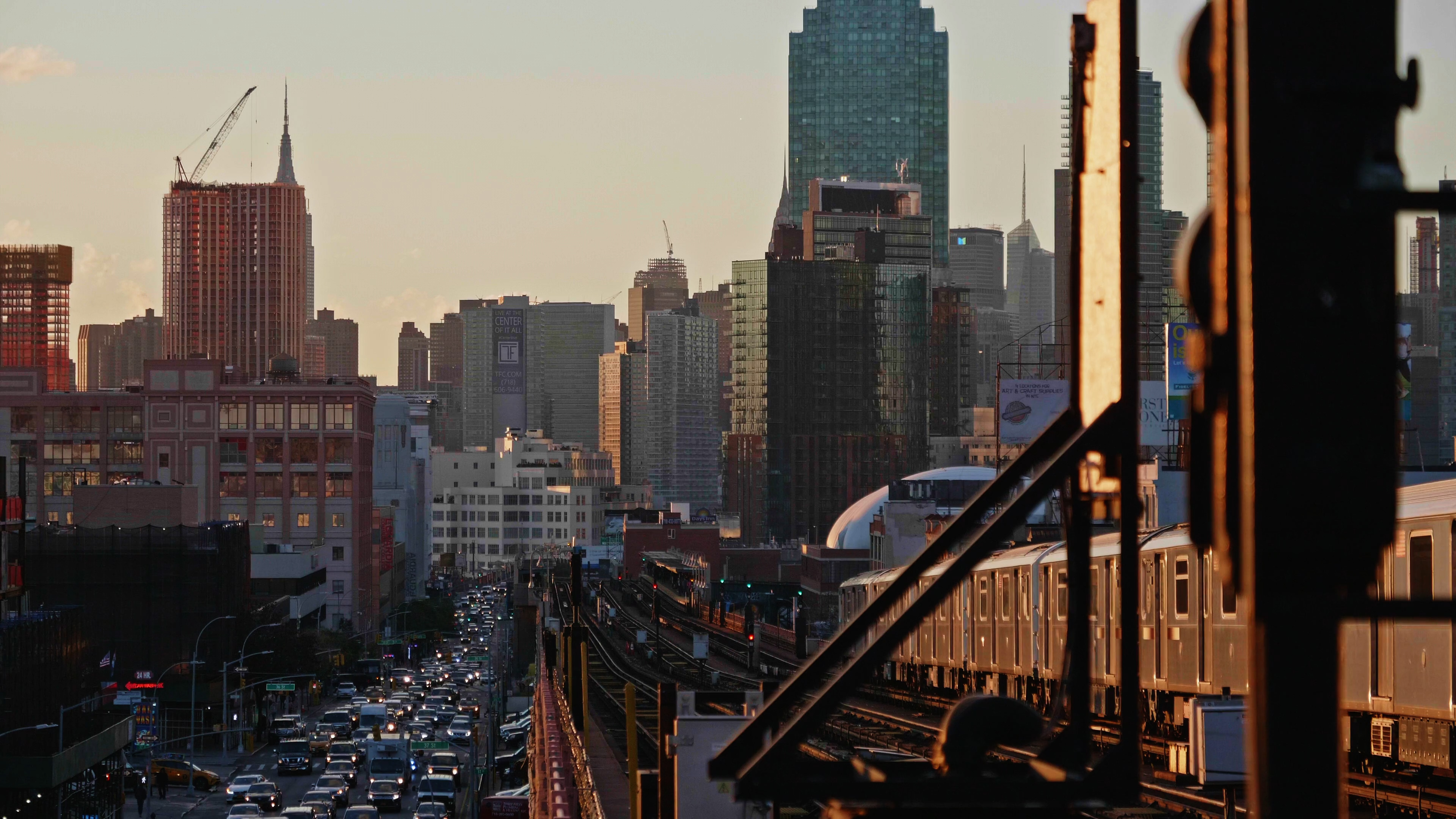 Sunlit subway train crossing a bridge with heavy traffic and skyscrapers in the background at dusk.