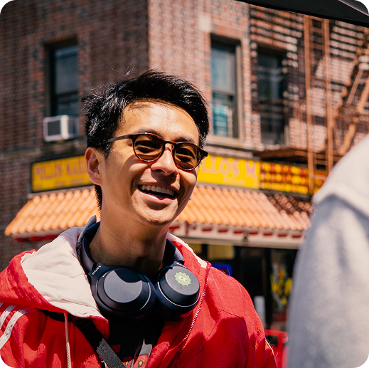 Smiling young man wearing sunglasses, red jacket, and black headphones around his neck standing outdoors in front of a building.