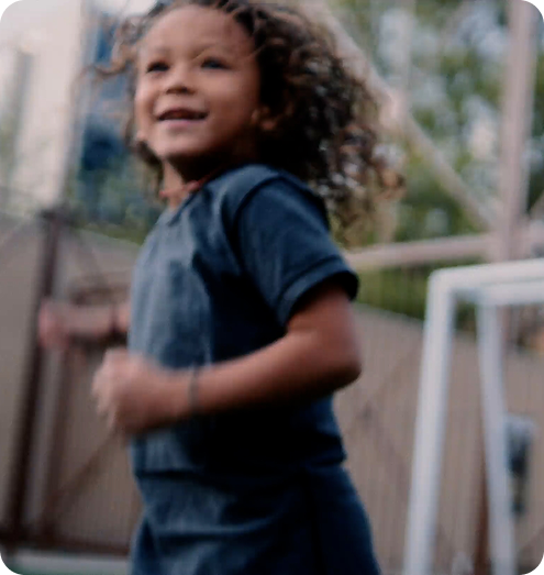 Smiling child with curly hair wearing a dark t-shirt playing outdoors near a soccer goal.