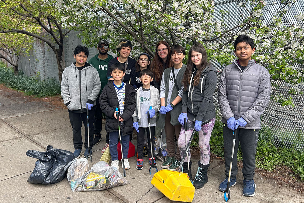 Group of diverse children and adults wearing gloves posing with litter pickers, a broom, and bags of collected trash on a sidewalk near blooming trees.
