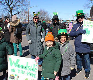 Group of people including children and adults wearing St. Patrick's Day themed hats and holding festive signs during an outdoor parade on a sunny day.