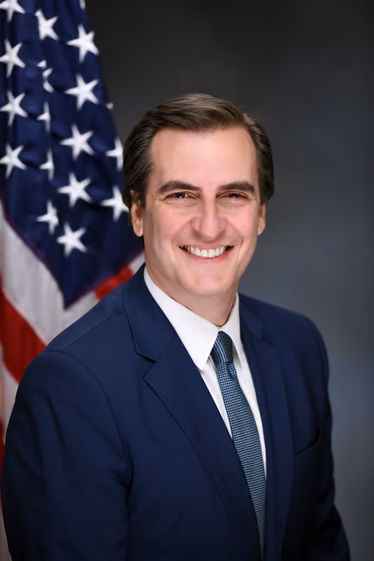 Man in blue suit and tie smiling in front of an American flag and dark background.