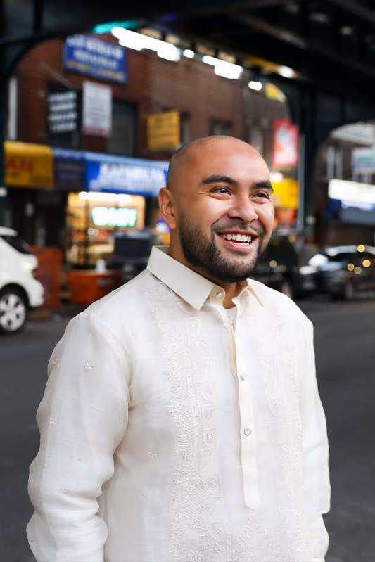 Smiling bald man with beard wearing a traditional embroidered white shirt outdoors in an urban setting.