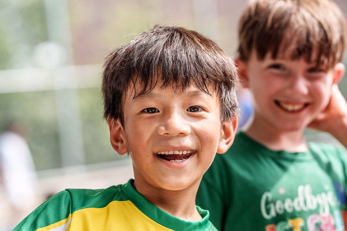 Two smiling boys outdoors, one wearing a yellow and green shirt, the other in a green shirt with text.