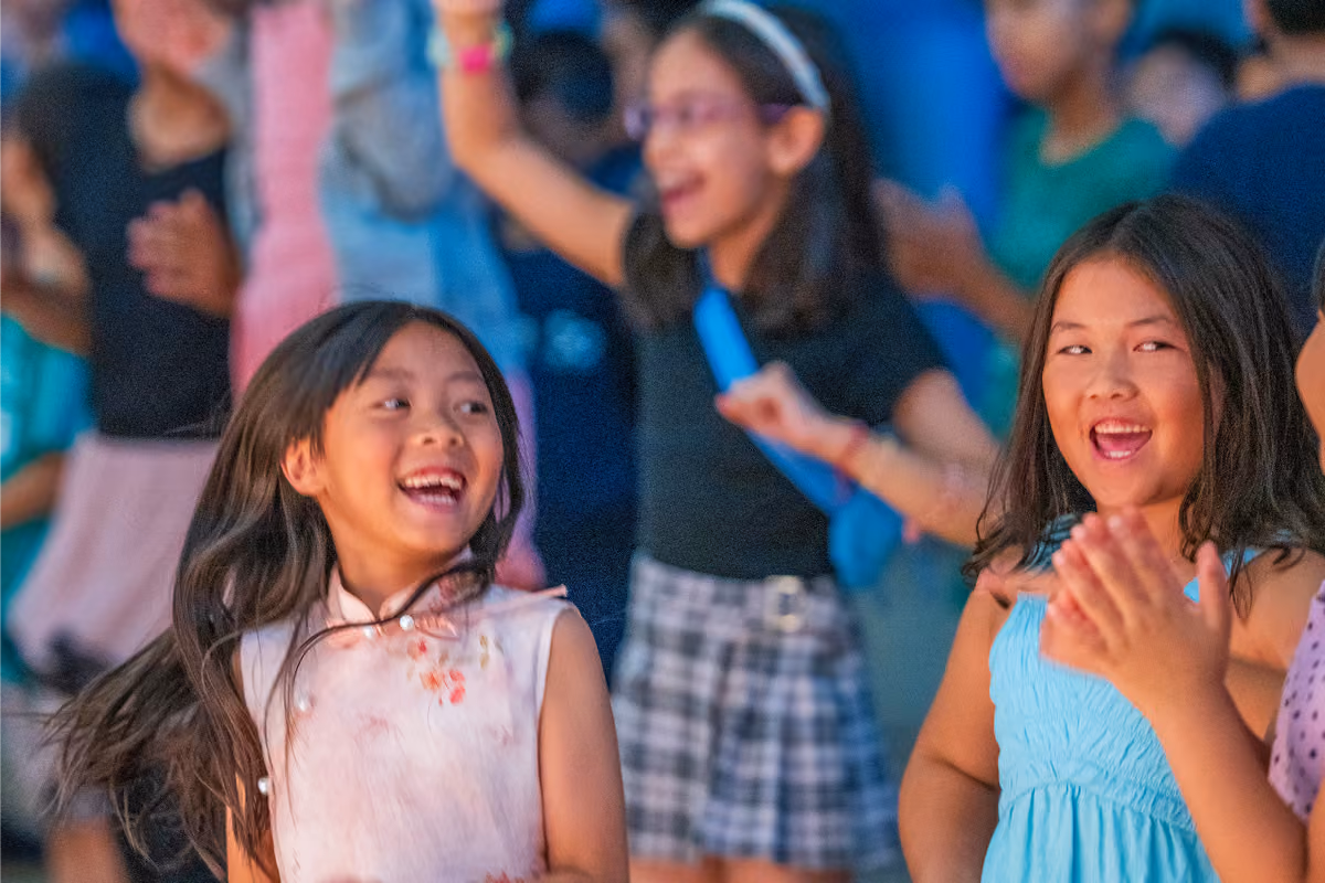 Two young girls laughing and clapping while others dance joyfully in the background.