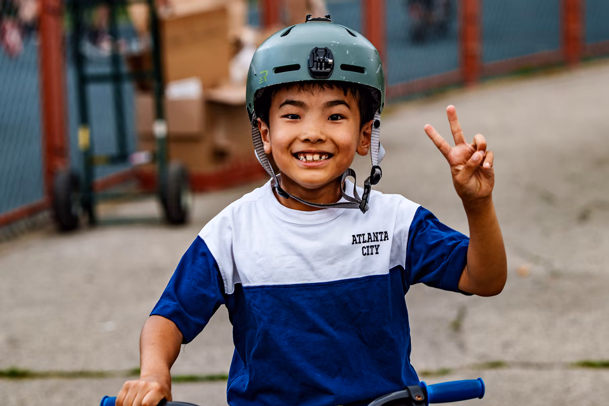 Smiling boy wearing helmet and Atlanta City shirt on a blue bike, showing peace sign with right hand.