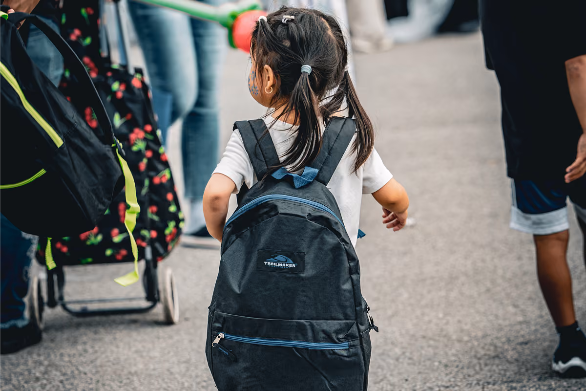 Young girl with pigtails wearing a backpack walking on a paved path among people.