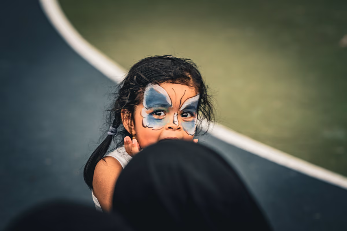Young girl with butterfly face paint looking over someone's shoulder on outdoor court.