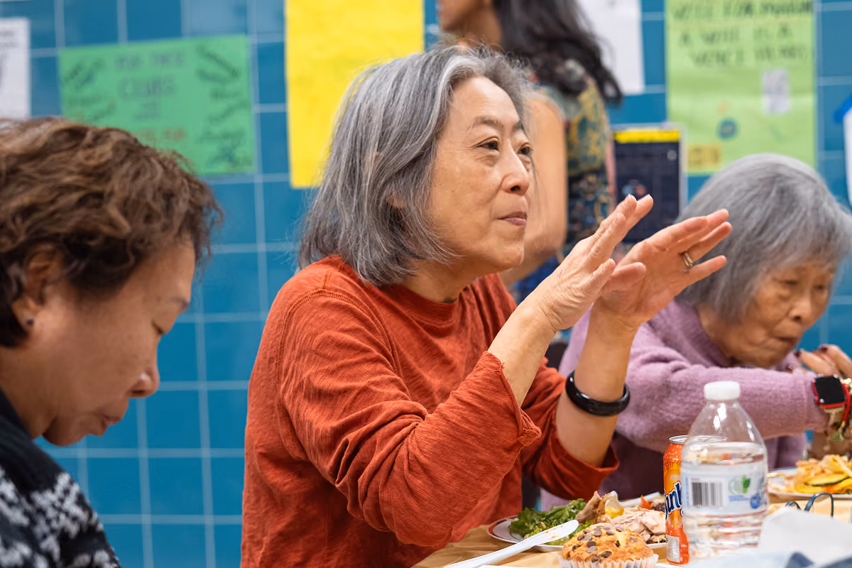 Three elderly women sitting at a table eating and talking, with drinks and food on the table.
