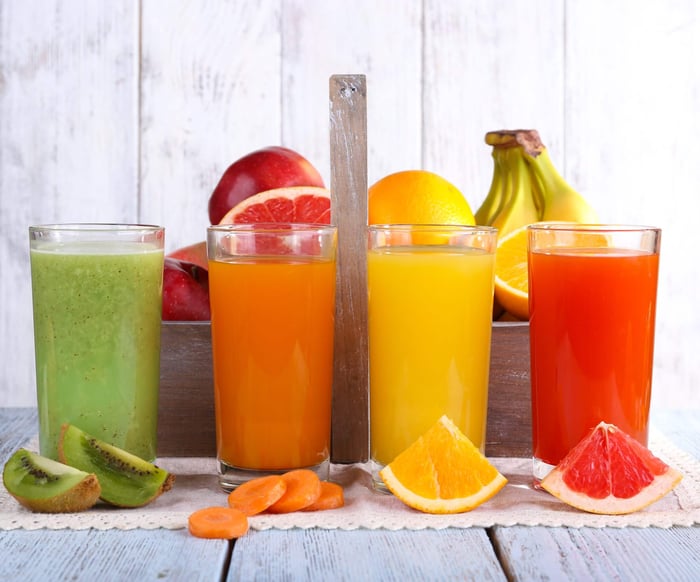 Three glasses of distinct types of juices surrounded by a variety of fruit.